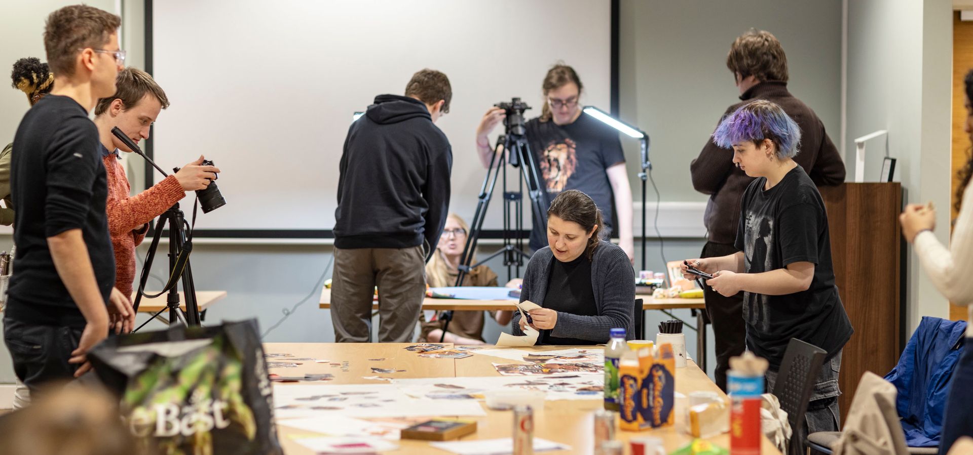 The Preservative Party working in a room with a large table. Some members are surrounding it cutting magazines, some are behind it filming