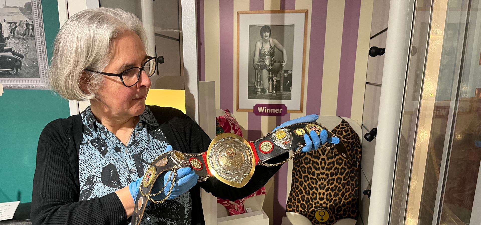 a curator holding Sue Brittain's wrestling belt in front of a wrestling display