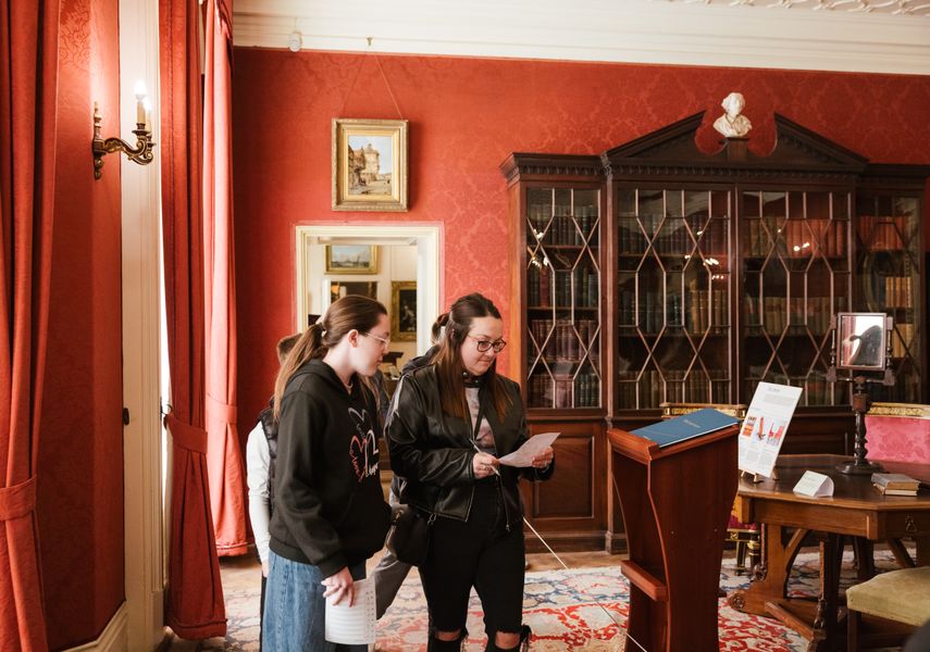 Two people looking at a trail sheet whilst looking around a red room with a large bookshelf