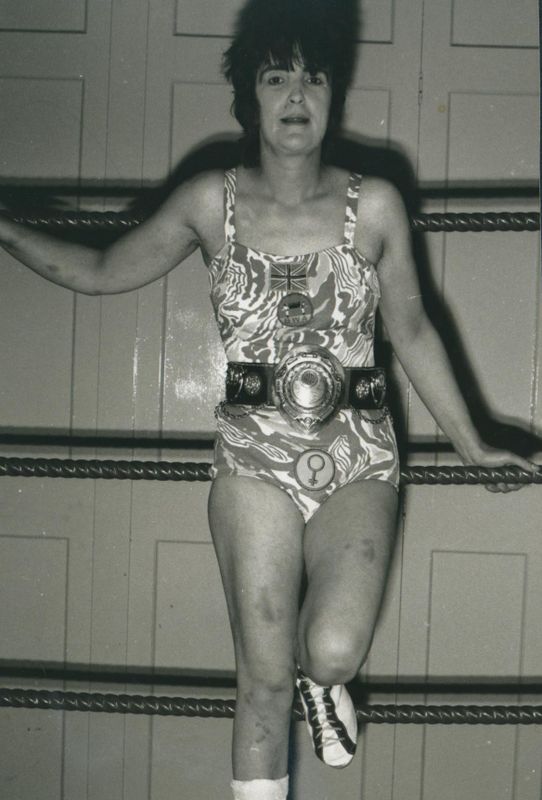 A black and white image of Sue Brittain in a costume and wrestling belt