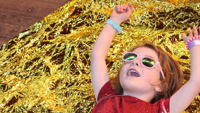 A toddler in sunglasses in a red sparkly dress laid on gold fabric
