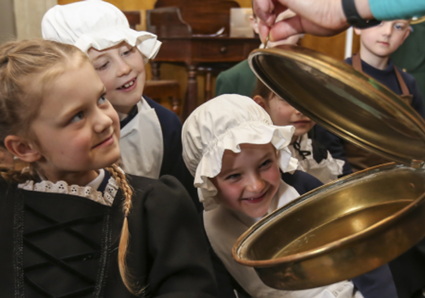 A group of children in traditional costumes looking at an old fashioned bed warmer.