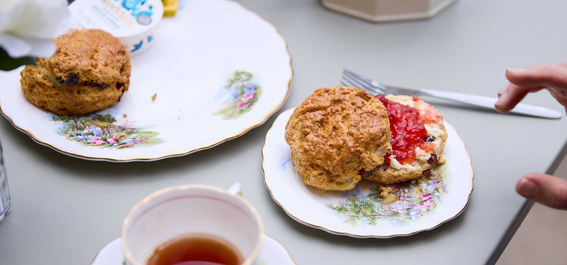 a hand reaching for a scone covered in jam and cream, with a cup of tea on the table an another plate with an extra scone on