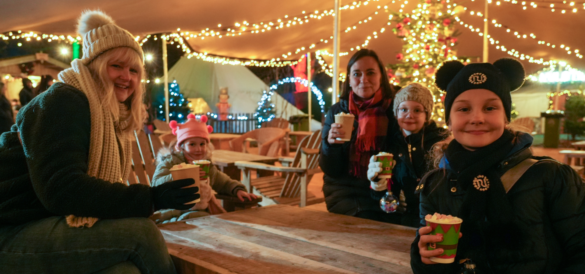 A family sat on a picnic bench under a tent canopy drinking hot chocolate