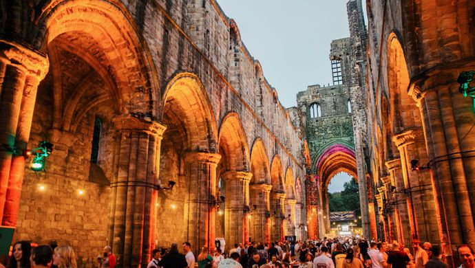 A Cistercian monastery lit up orange at dusk with lots of people enjoying beer and food.