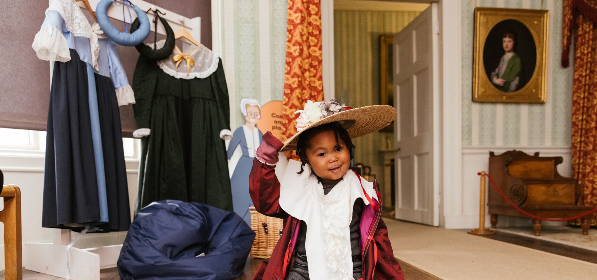 A toddler kneeling on the floor in fancy dress wearing a hat