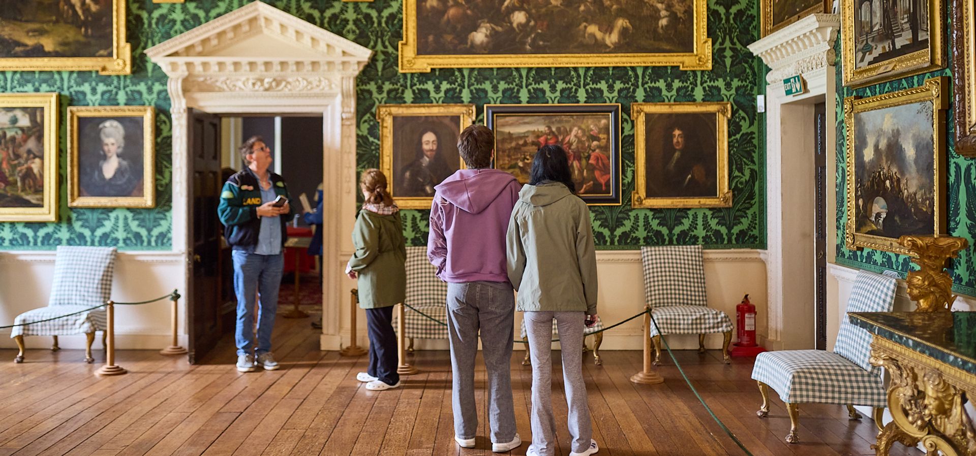 A group of people looking around a room in a heritage house with green wallpaper and ornate framed works on the wall