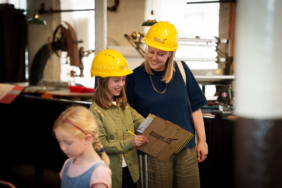 Two children with an adult looking at a clipboard, all wearing yellow hard hats
