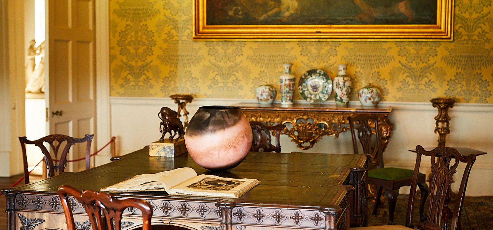 A wallpapered room with a large 4 person desk, decorative items and a painting on the wall