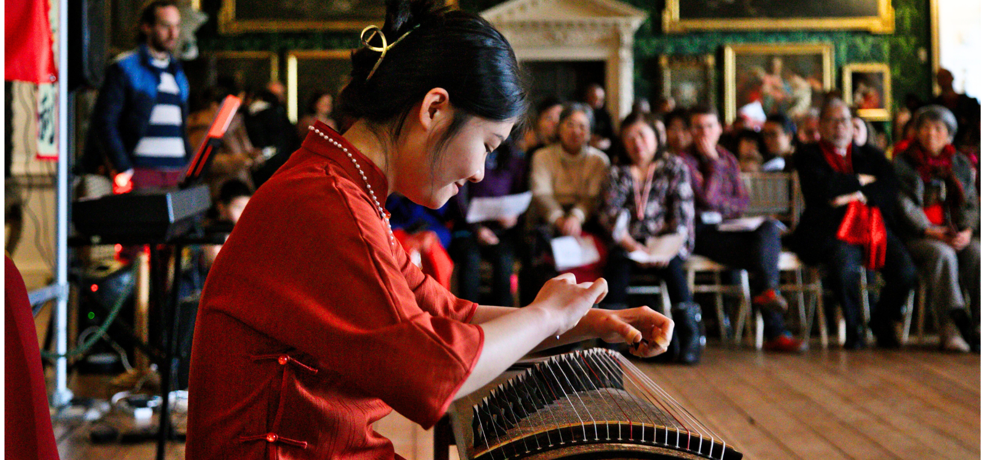 A woman banging on a drum in a red top, surrounded by people sat down in Temple Newsam