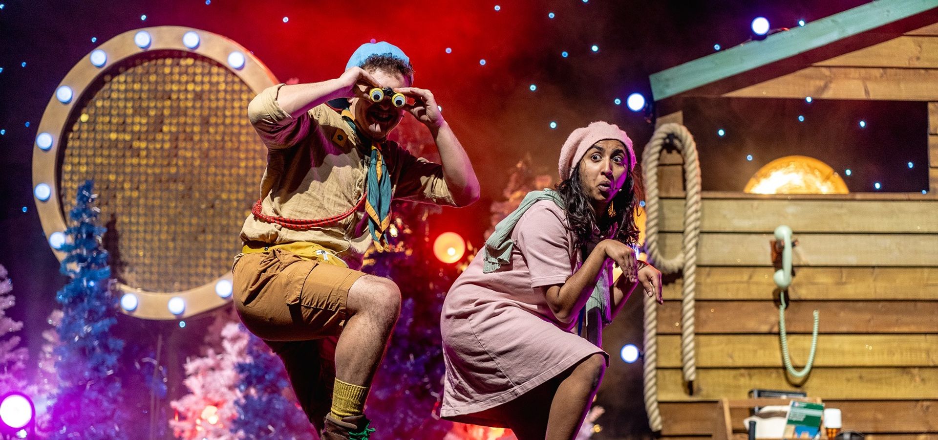 A man and a woman in costume on stage, both standing on one leg. The man is using binoculars